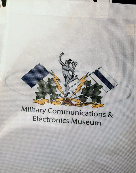 A white reusable tote bag printed with the logo and text for the Military Communications & Electronics Museum, featuring a central emblem with flags, lightning bolts, and a banner.