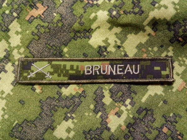 Cadpat name tag with green embroidered edges and two swords crossed in white embroidery, the name Bruno is also embroidered in the same colour. The name tag is sitting on top of a cadpat background.